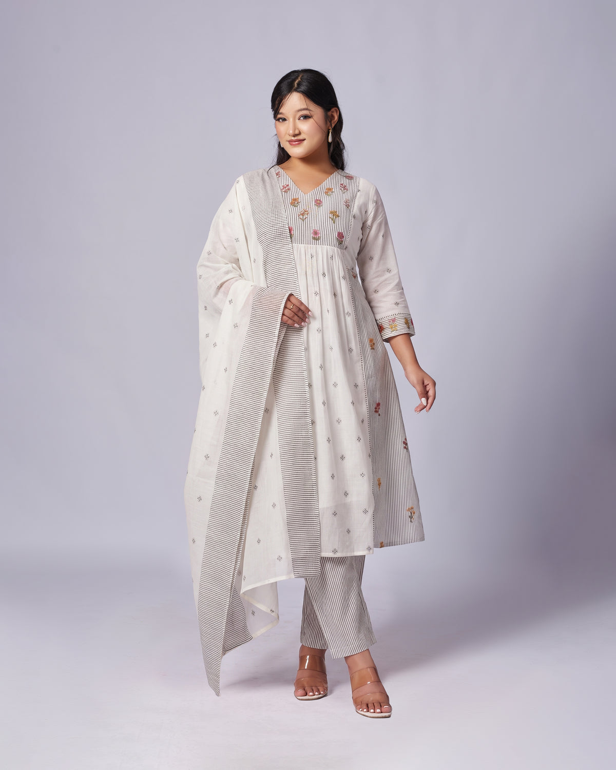 A woman modeling a traditional Indian outfit consisting of a light beige kurta with colorful floral embroidery, matching trousers, and a coordinating dupatta, set against a plain light gray studio background.