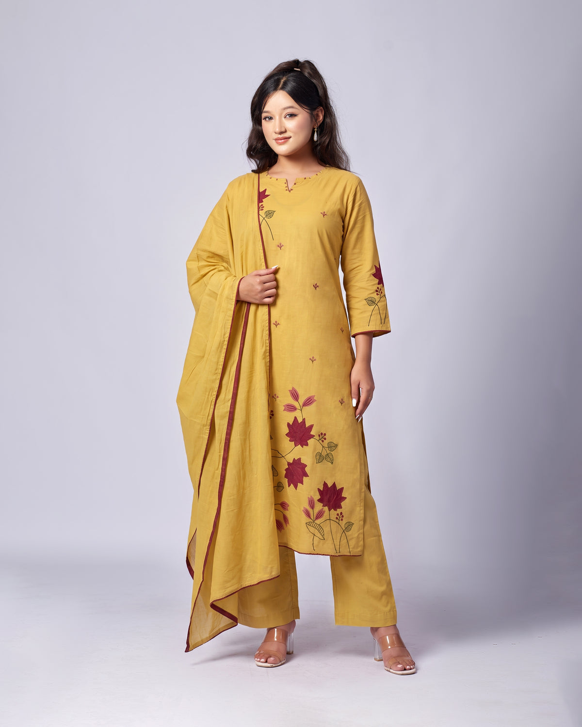 A woman modeling a mustard yellow ethnic suit set with floral embroidery and a matching dupatta, photographed against a light gray studio background.