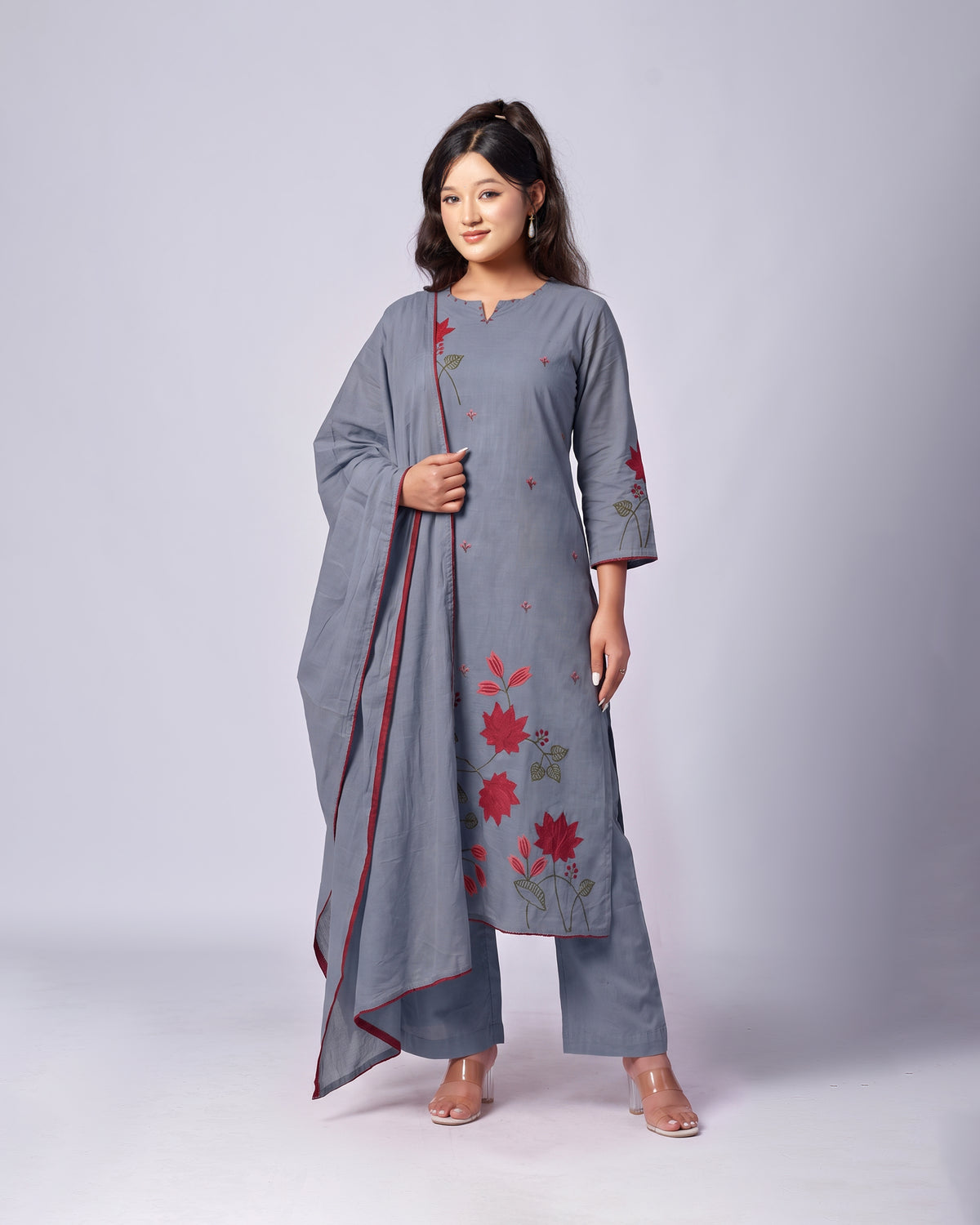 A woman modeling a gray three-piece ethnic suit set featuring a kurta with red floral embroidery, matching wide-leg trousers, and a coordinating dupatta with red border detailing, set against a plain light gray studio background.