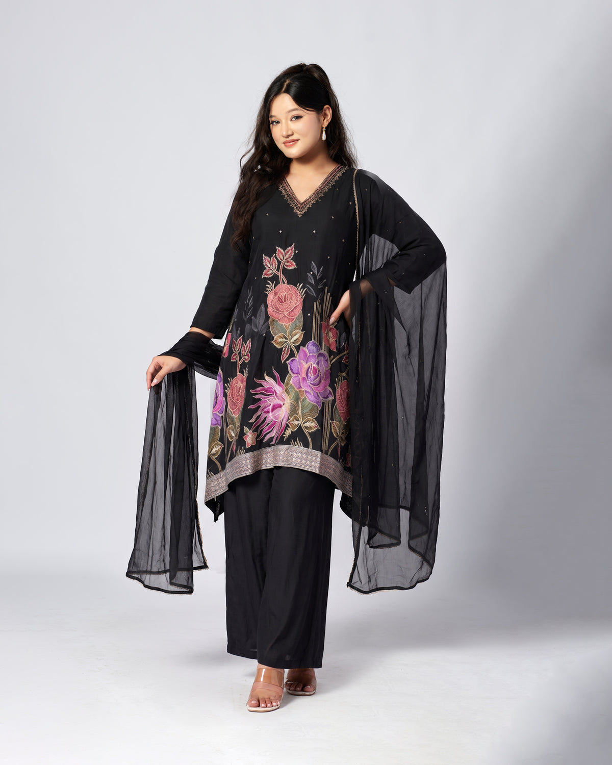 A woman modeling a black three-piece ethnic suit set with a floral embroidered kurta, black wide-leg pants, and a sheer black dupatta, photographed against a light gray studio background.
