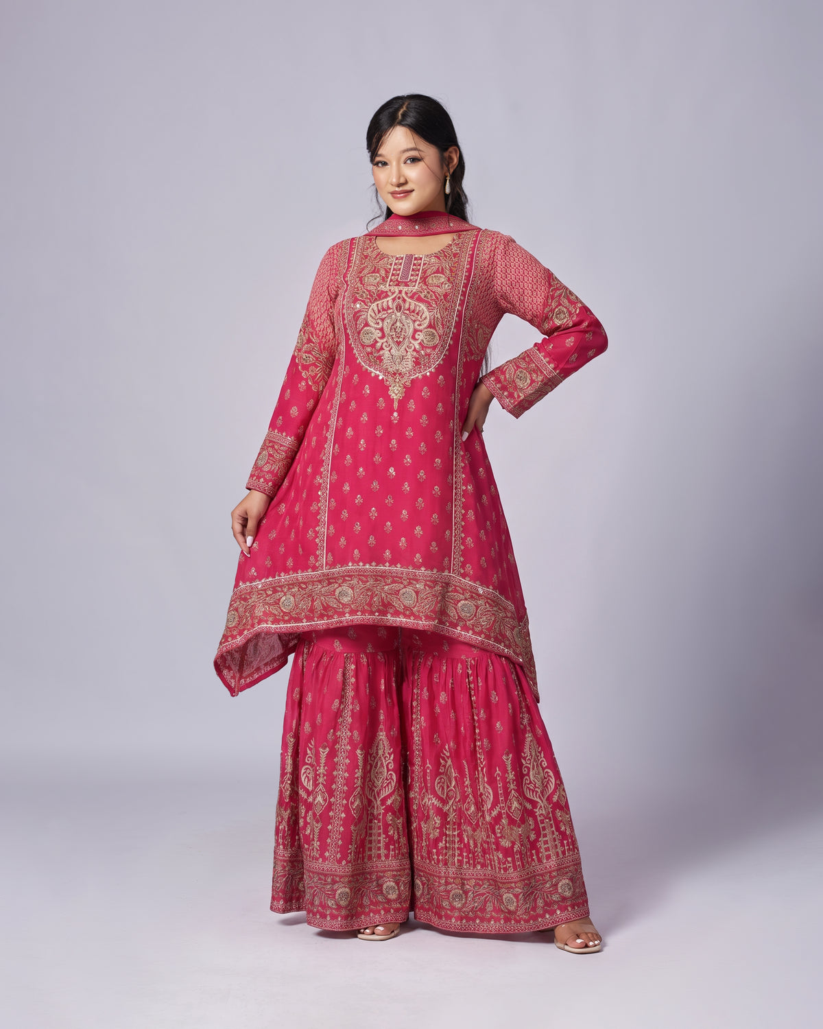 A woman modeling a traditional Indian outfit consisting of a pink kurta with gold embroidery, matching flared pants, and a dupatta, set against a plain light gray studio background.