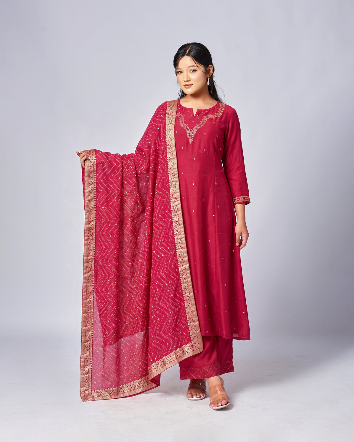 A woman modeling a traditional Indian salwar kameez set in deep magenta pink, featuring a kurta with gold embroidery, matching pants, and a sheer patterned dupatta with a gold border, set against a plain light gray studio background.
