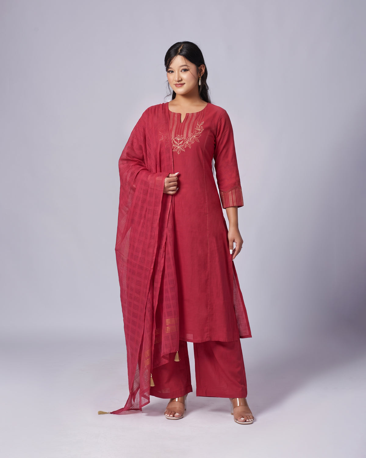 A woman modeling a three-piece traditional Indian salwar kameez suit in a deep red color, featuring a straight kurta with gold embroidery on the chest, matching wide-leg trousers, and a coordinating dupatta, set against a plain light gray studio background.