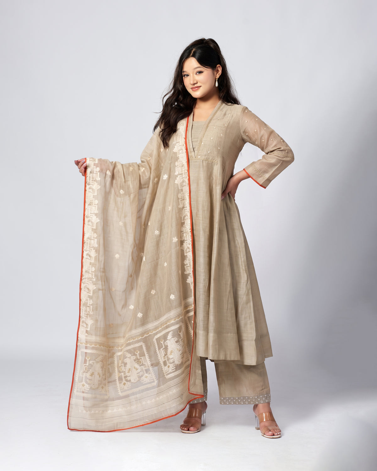 A woman modeling a traditional beige salwar kameez suit with matching pants and a sheer dupatta featuring white floral prints and a red-orange border, set against a light gray studio background.