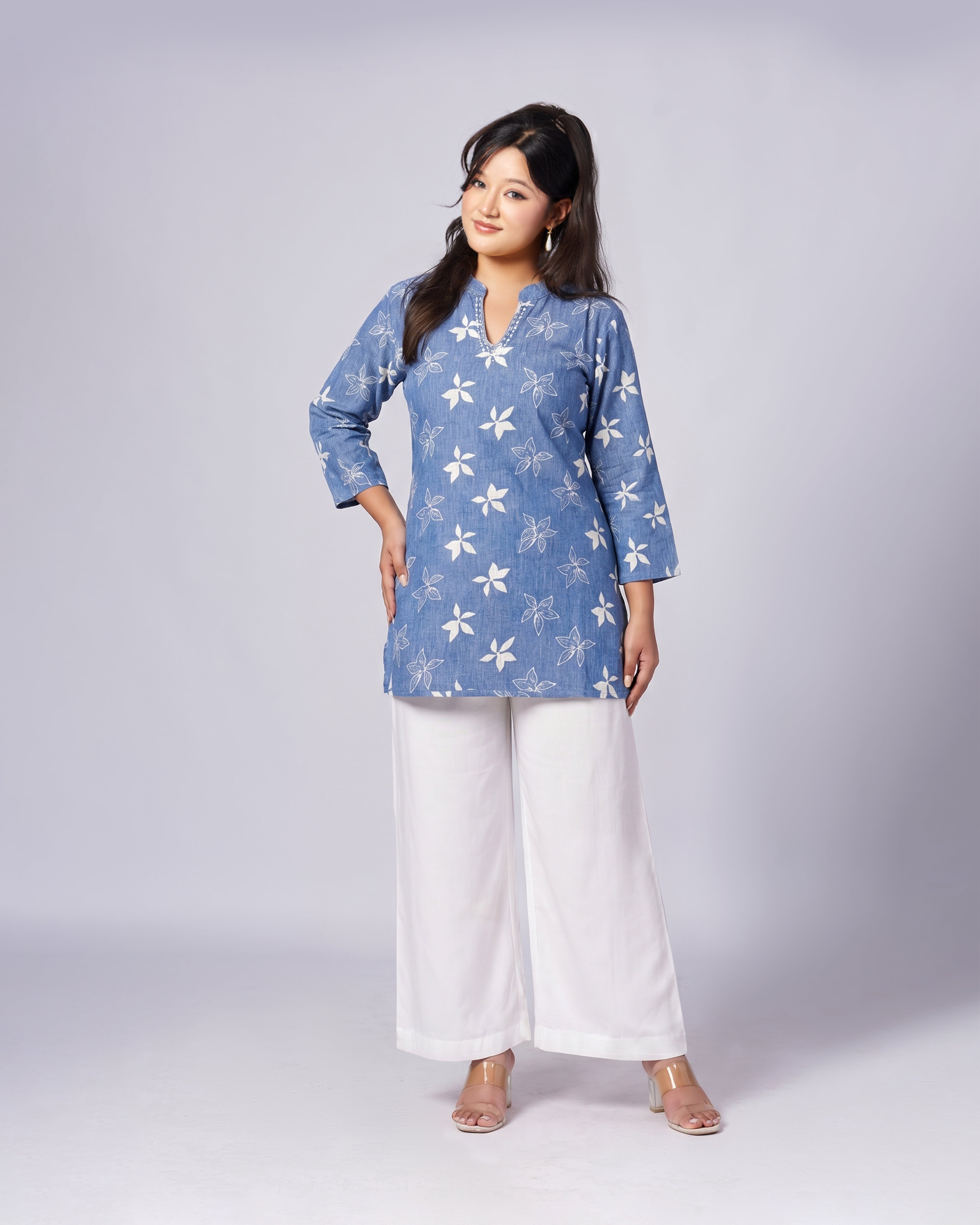 A woman modeling a light blue long-sleeve tunic top with a white floral pattern, paired with white wide-leg pants, against a light gray studio background.