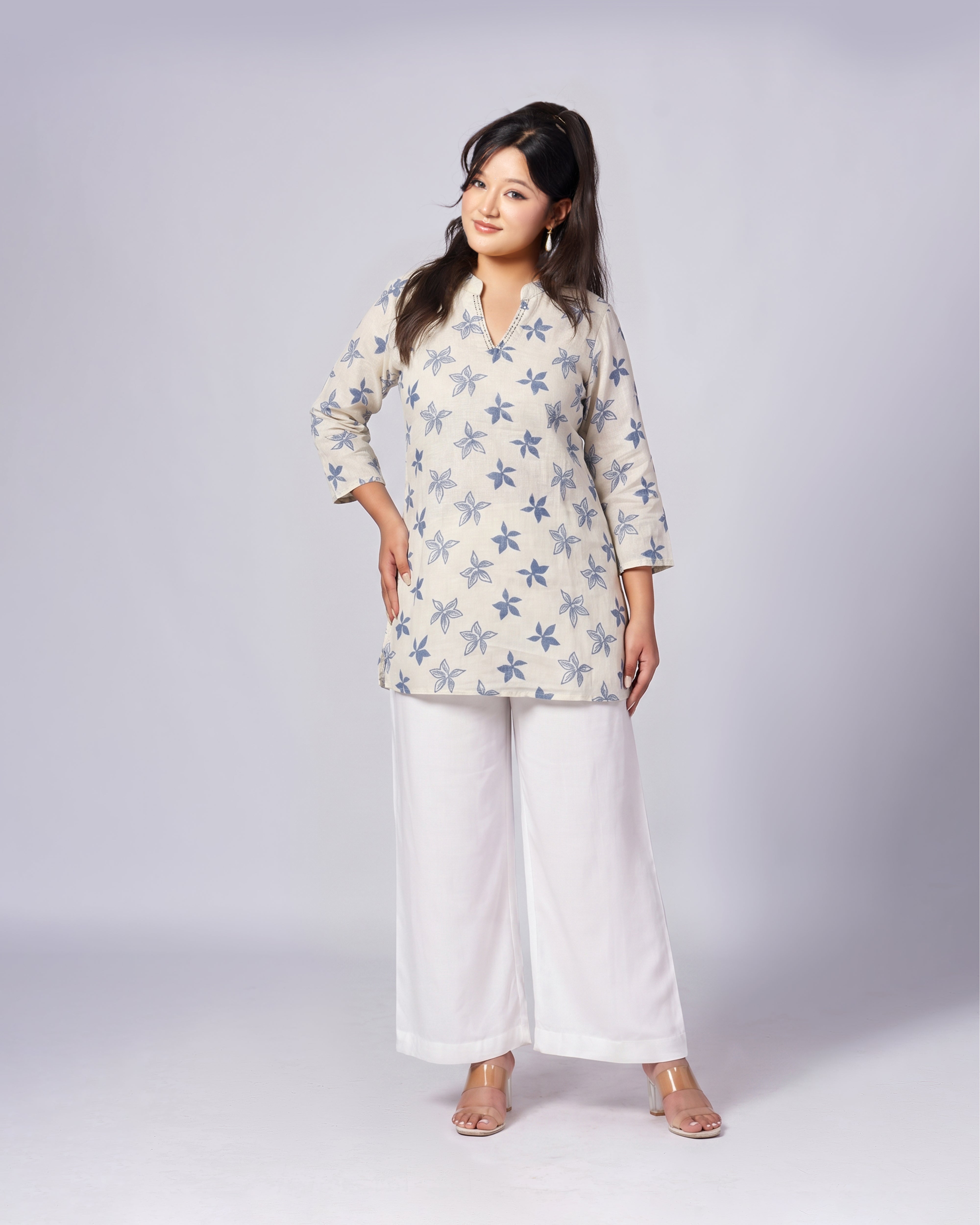 A woman modeling a light beige kurta-style top with a blue star pattern, paired with white wide-leg pants, against a light gray studio background.