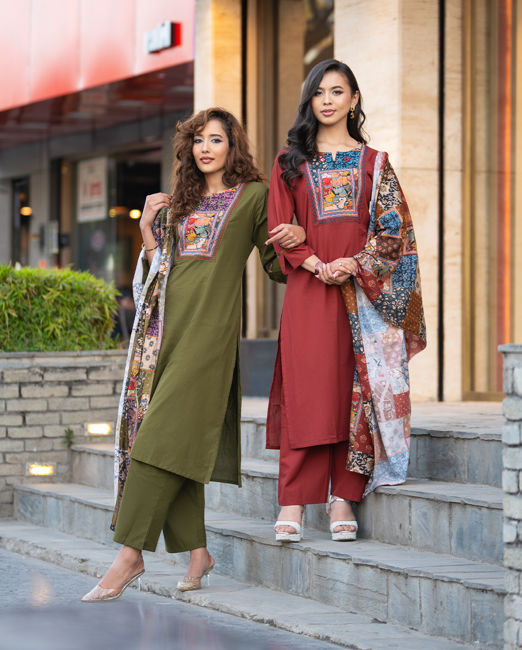 Two women in traditional outfits with embroidered shawls standing on steps outdoors.