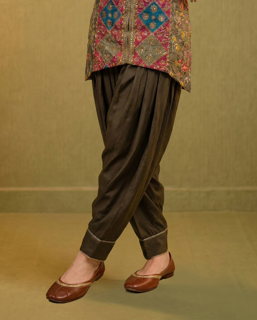 Person wearing a traditional outfit with embroidered top and brown pants on a beige background