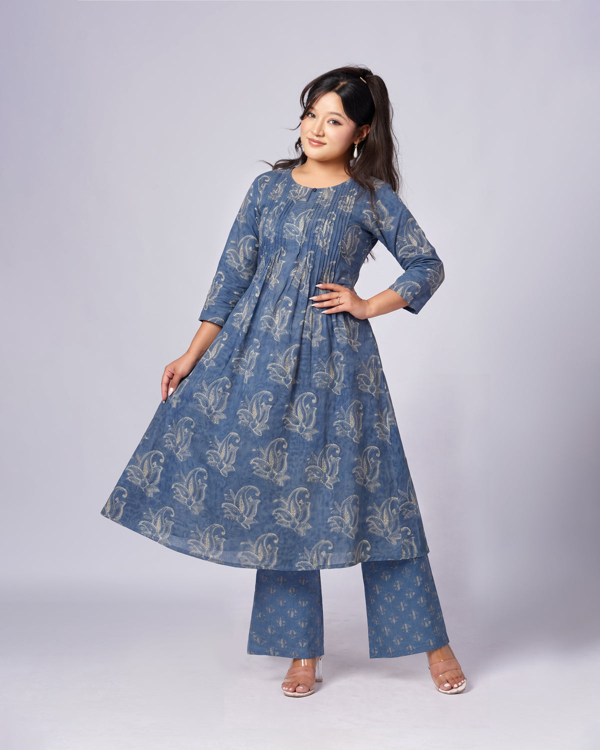 A woman modeling a blue three-piece Indian ethnic wear set, featuring a long-sleeve kurta with a beige floral block print, matching wide-leg trousers, and a coordinating belt, set against a plain light gray studio background.