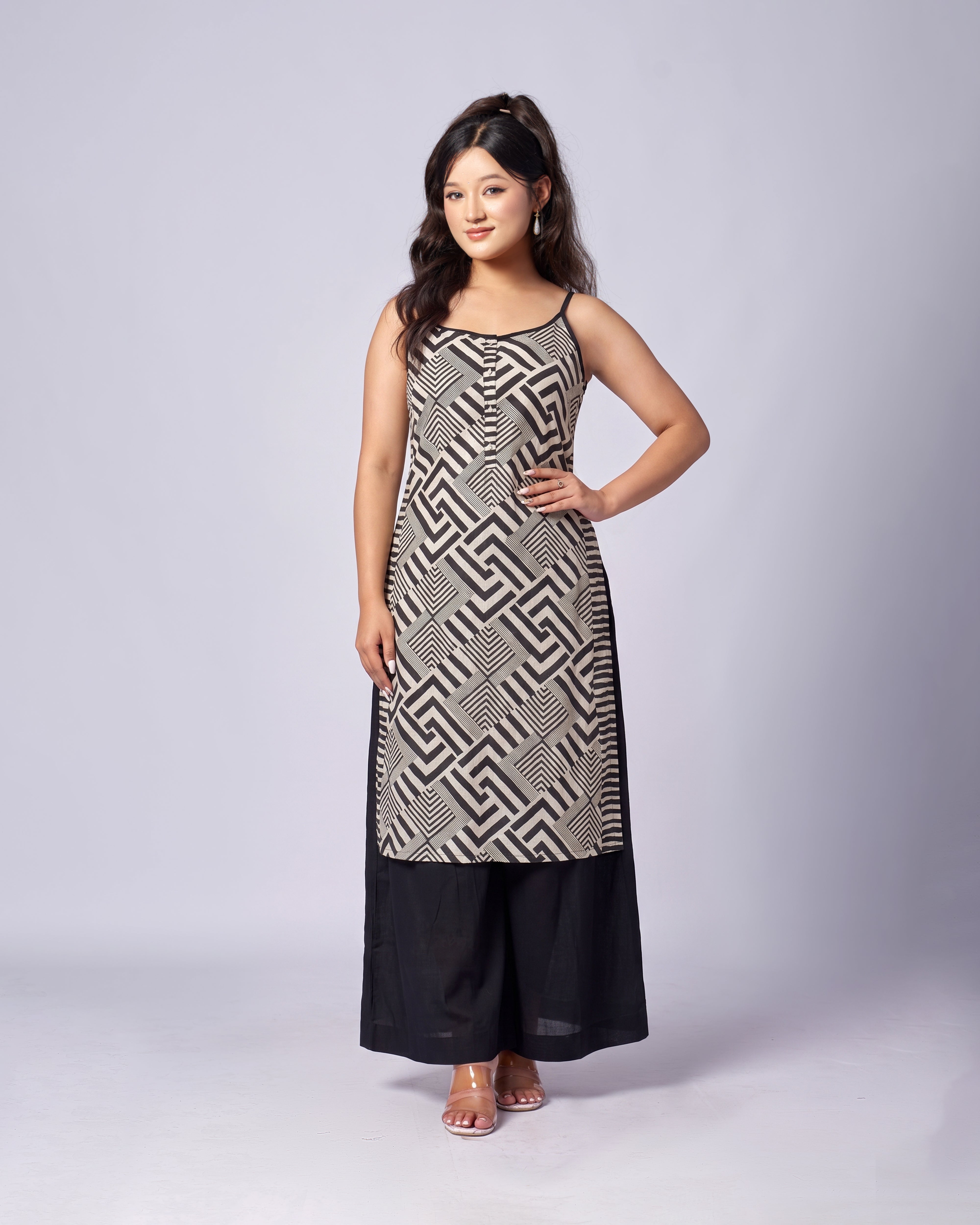 A woman modeling a sleeveless black and white geometric patterned tunic dress with black wide-leg pants, standing against a light gray studio background.
