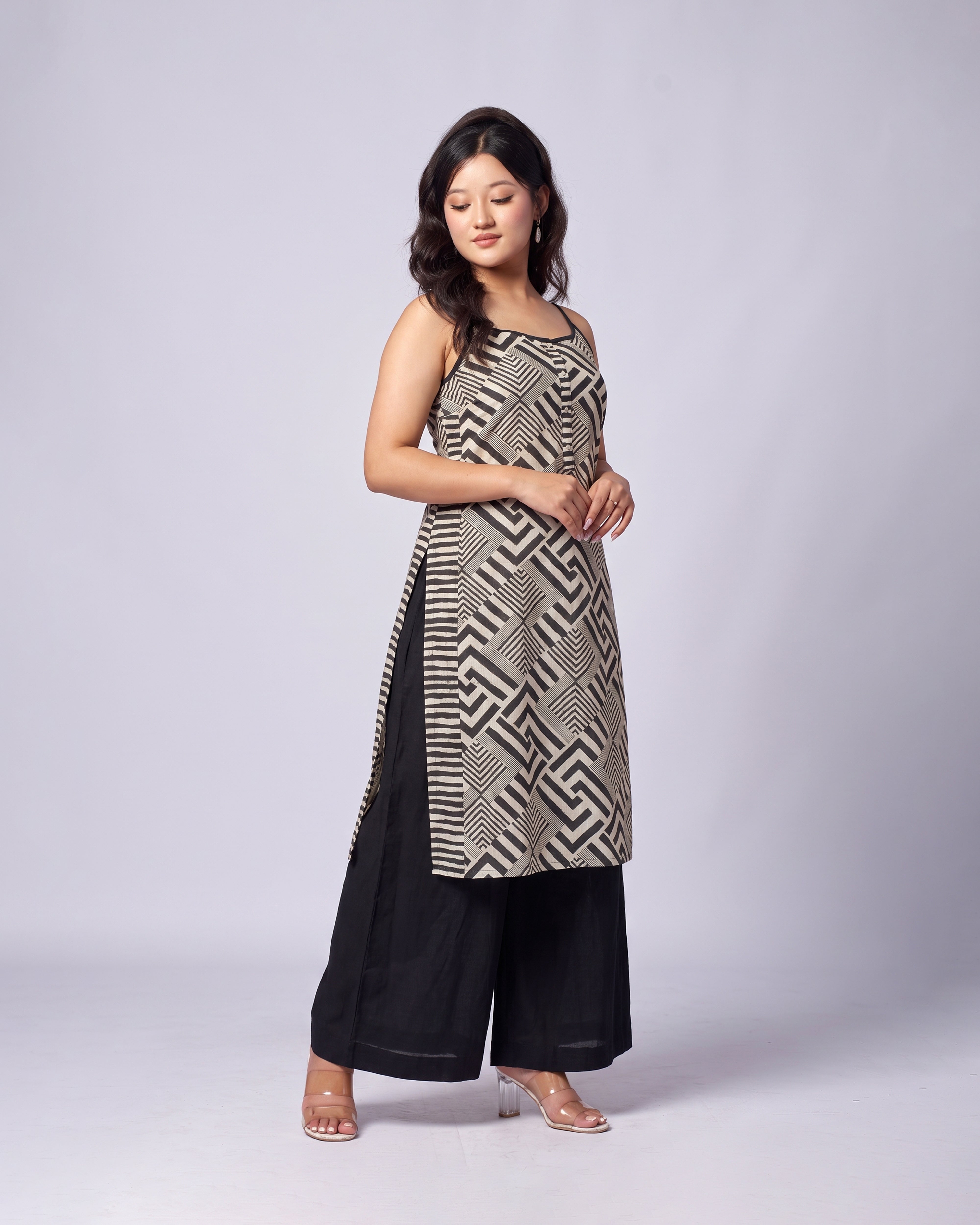 A woman modeling a sleeveless black and beige geometric patterned tunic with black wide-leg pants, standing against a light gray studio background.