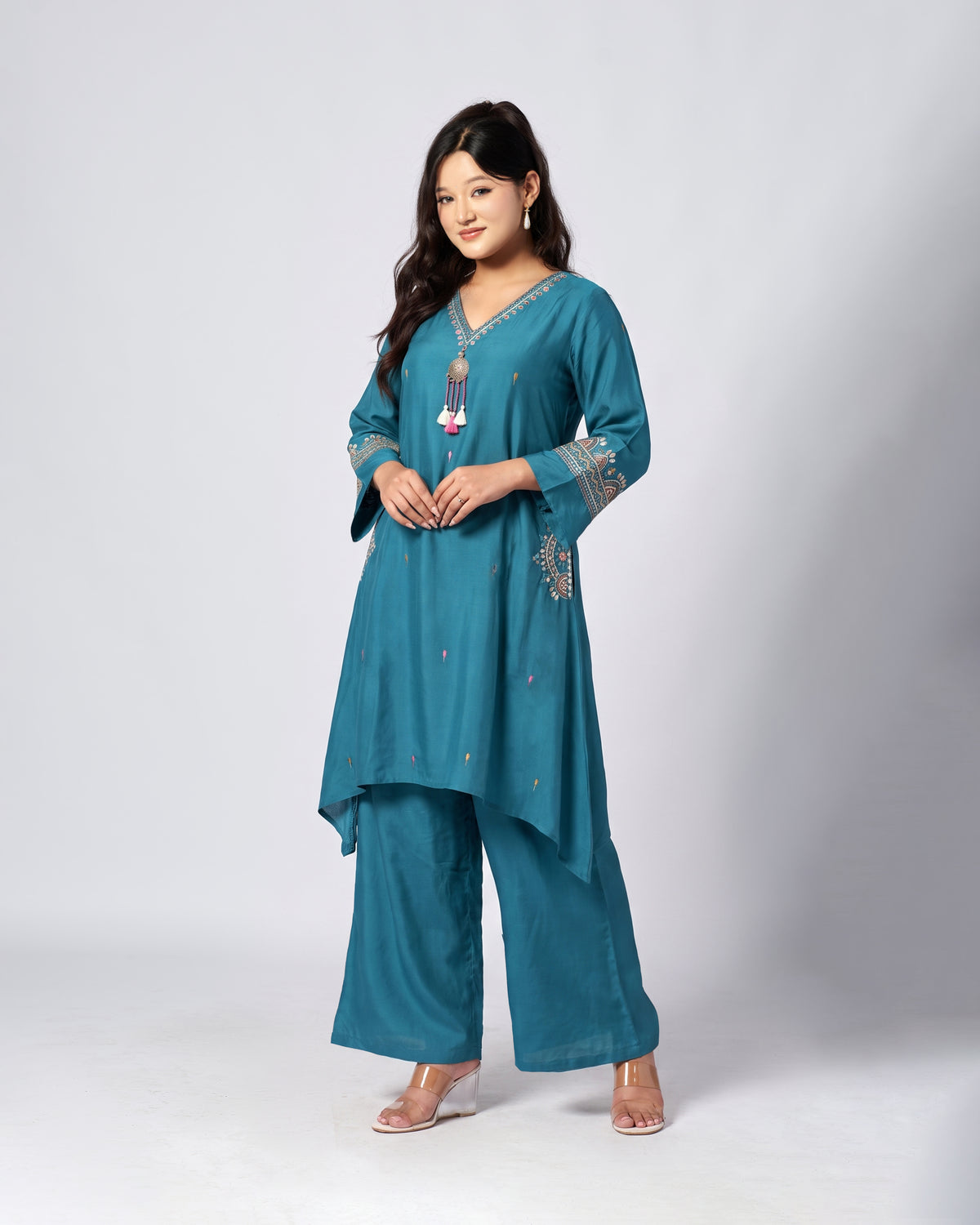 A woman modeling a teal blue-green traditional Indian outfit consisting of a long kurta with embroidered details and matching wide-leg pants, photographed against a light gray studio background.