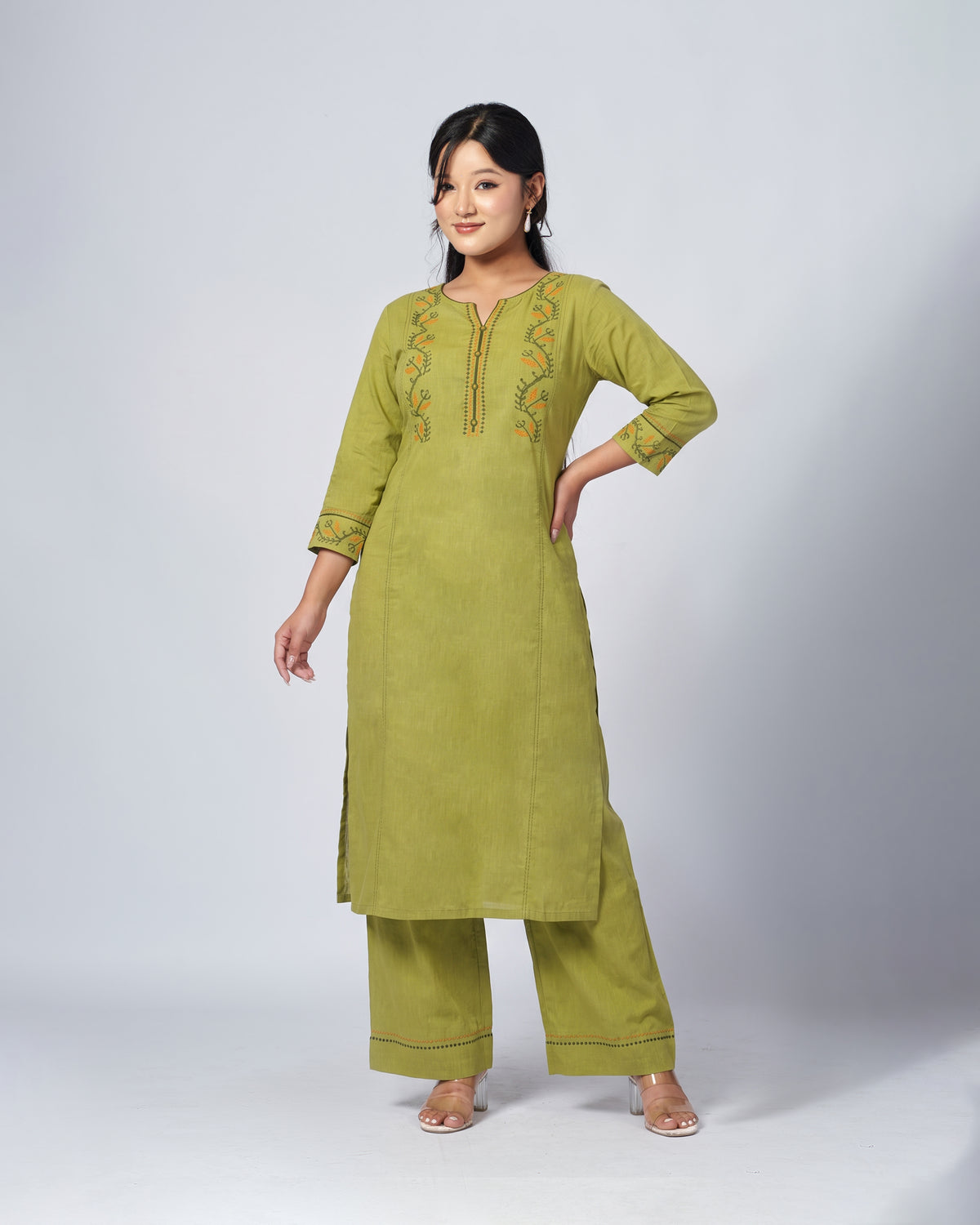 A woman modeling a three-piece olive green kurta set with matching wide-leg trousers, featuring colorful embroidered details on the chest and cuffs, set against a plain light gray studio background.