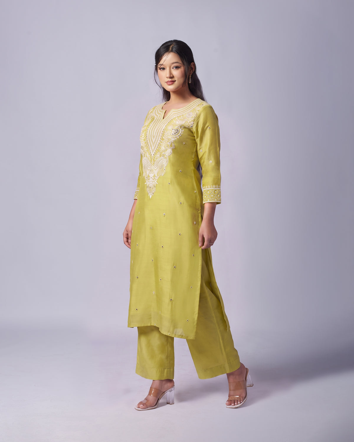 A woman modeling a light yellow-green kurta set with matching pants, featuring white embroidered detailing on the chest and cuffs, set against a plain light gray studio background.