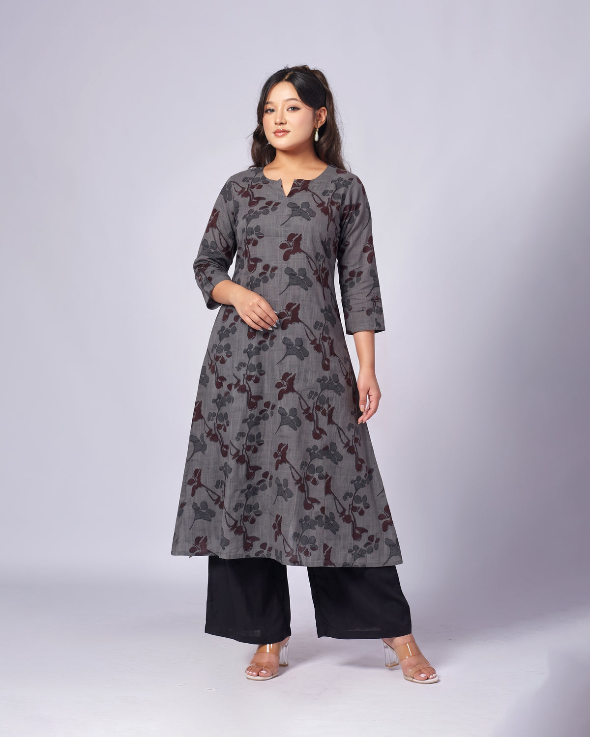 A woman modeling a long gray tunic with a dark floral pattern, paired with black wide-leg pants and clear high-heeled sandals, against a light gray studio background.