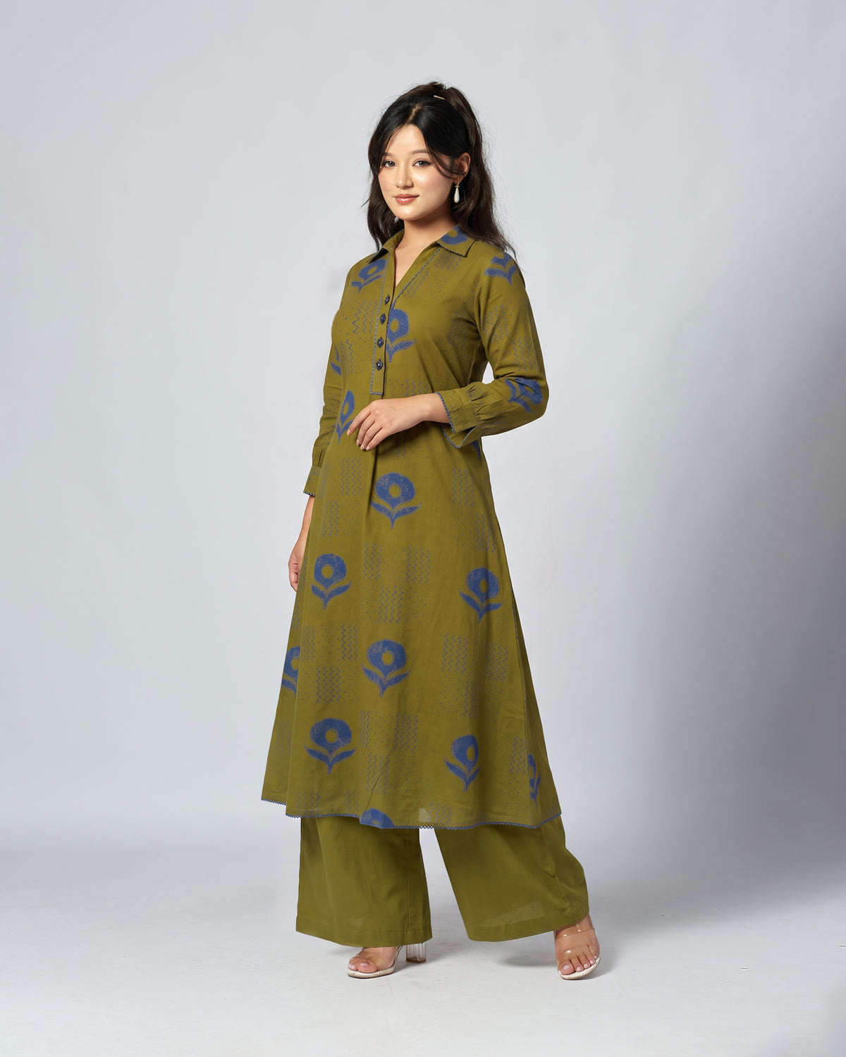 A woman modeling a two-piece olive green kurta and trouser set with blue floral block prints, photographed against a neutral light gray studio background.