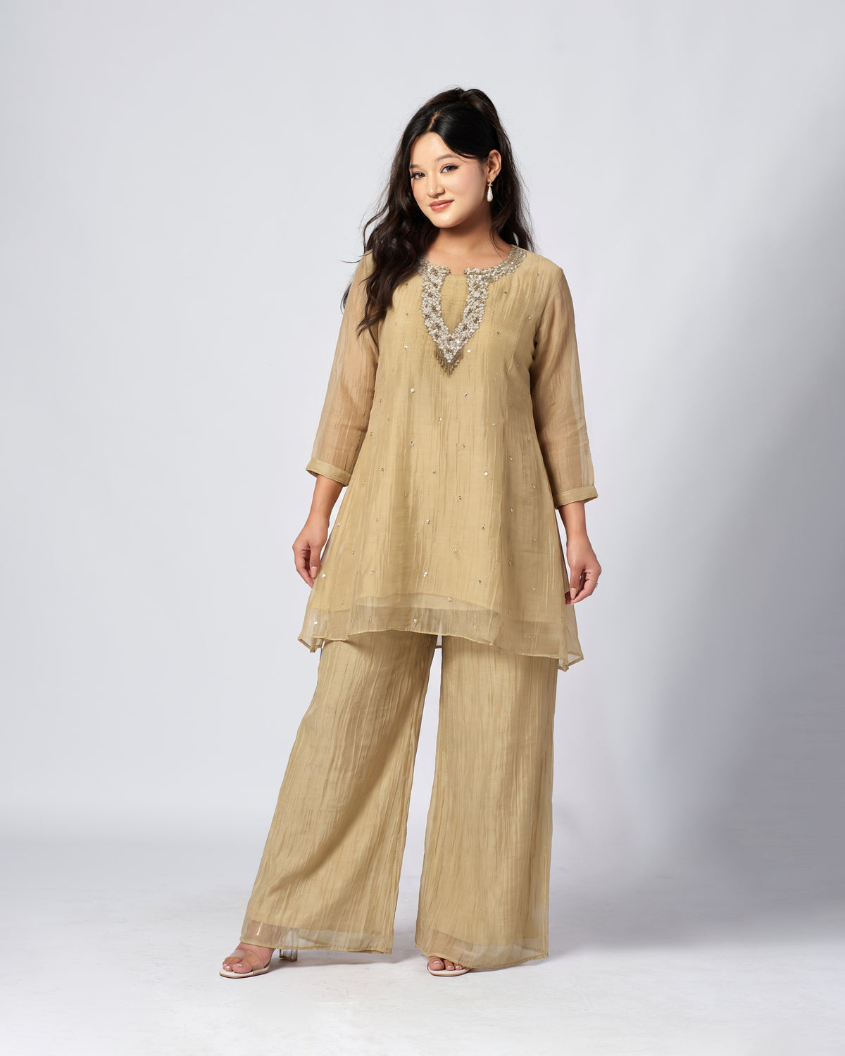 A woman modeling a beige three-piece kurta set with matching wide-leg pants and a sheer embroidered V-neckline detail, photographed against a light gray studio background.