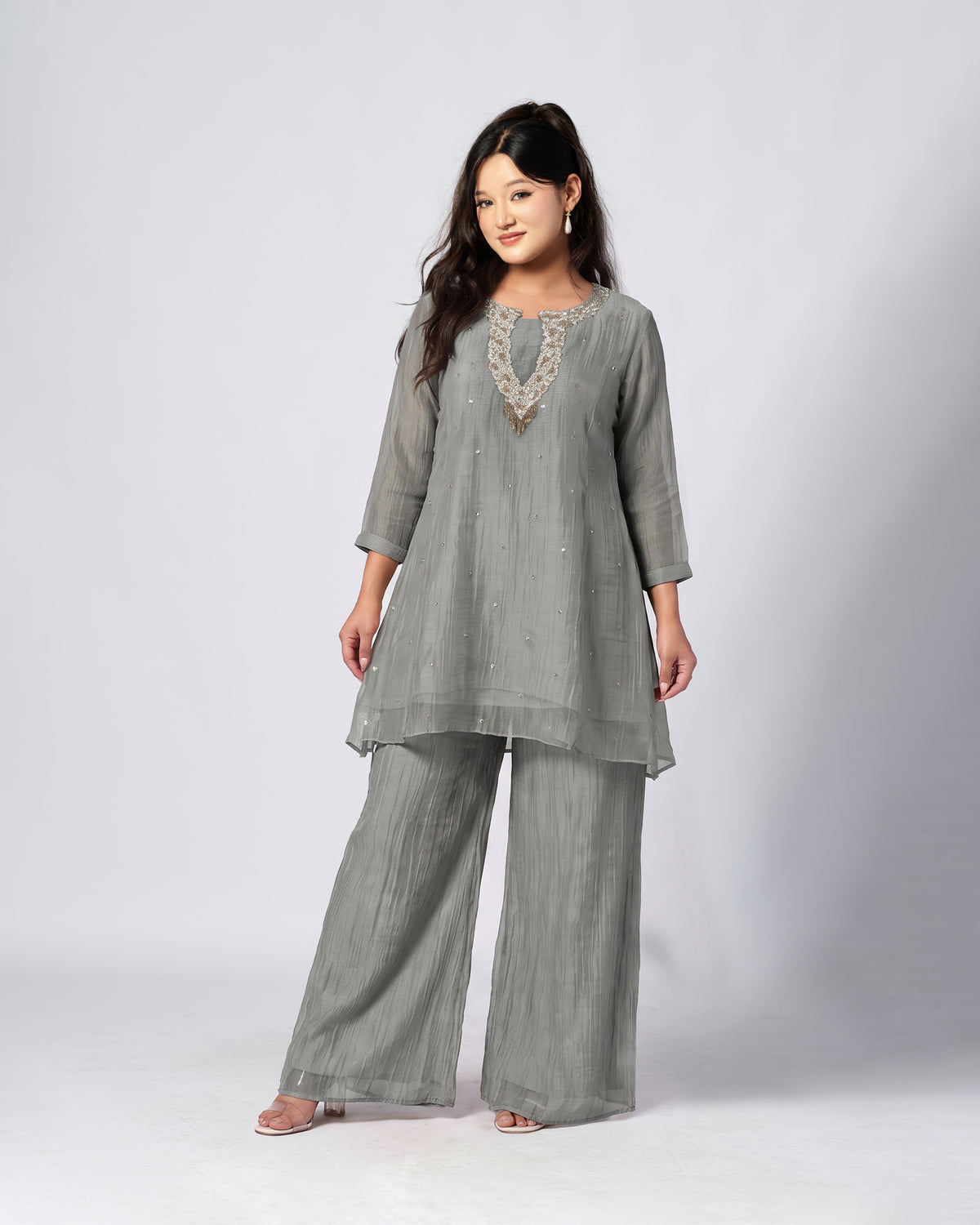 A woman modeling a Grey three-piece kurta set with matching wide-leg pants and a sheer embroidered V-neckline detail, photographed against a light gray studio background.