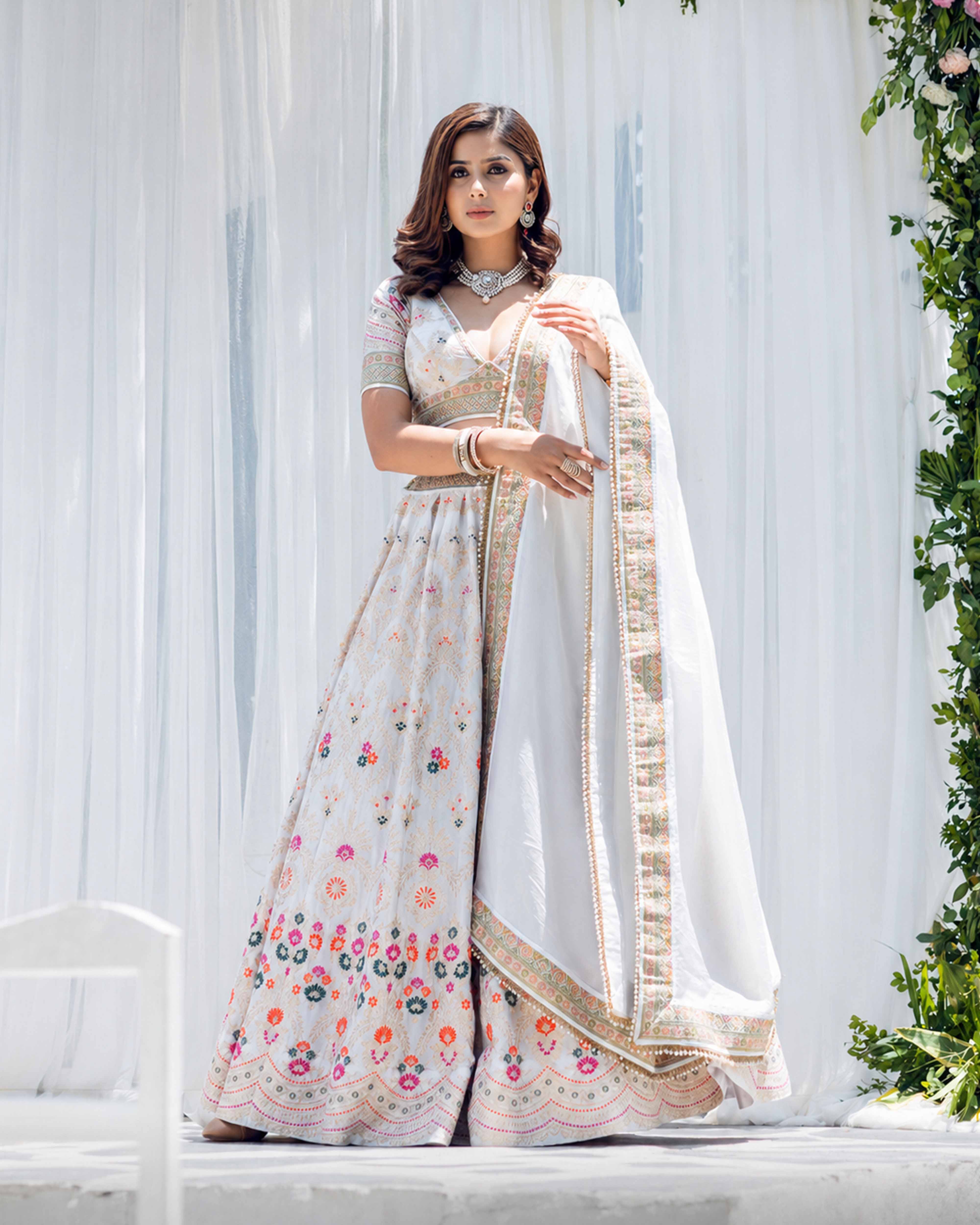Woman in a white saree with floral patterns standing against a white curtain.