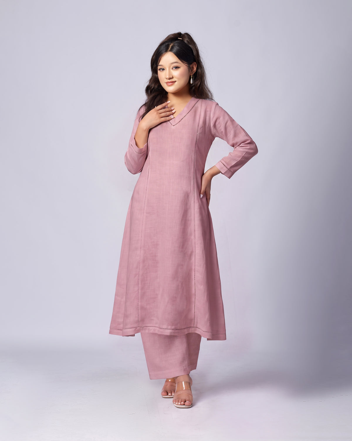 A woman modeling a dusty pink long kurta with matching pants and clear high-heeled sandals, standing against a light gray studio background.