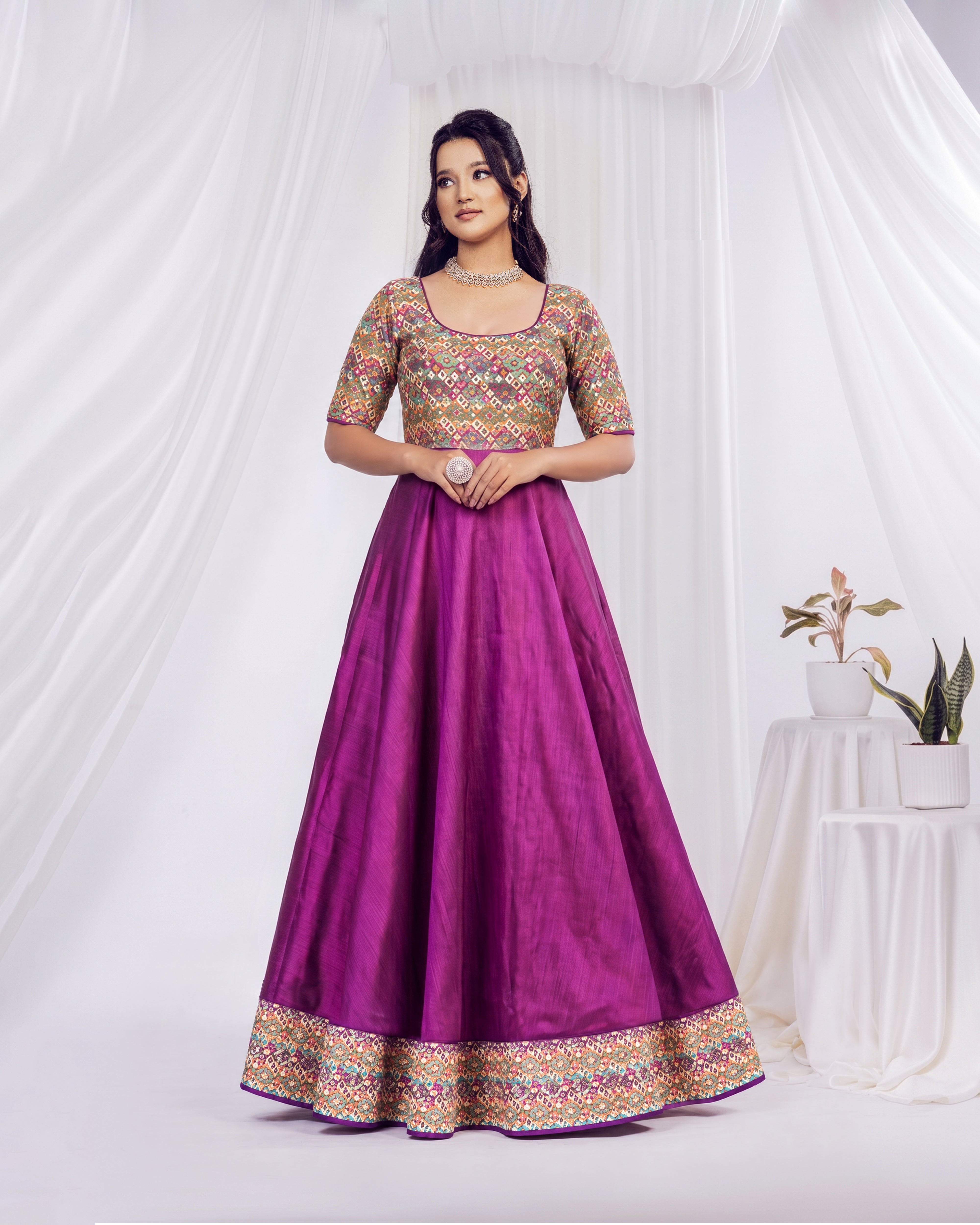 A woman models a traditional Indian Anarkali gown featuring a vibrant purple skirt and a heavily embroidered gold, pink, and green blouse, set against a draped white fabric backdrop.