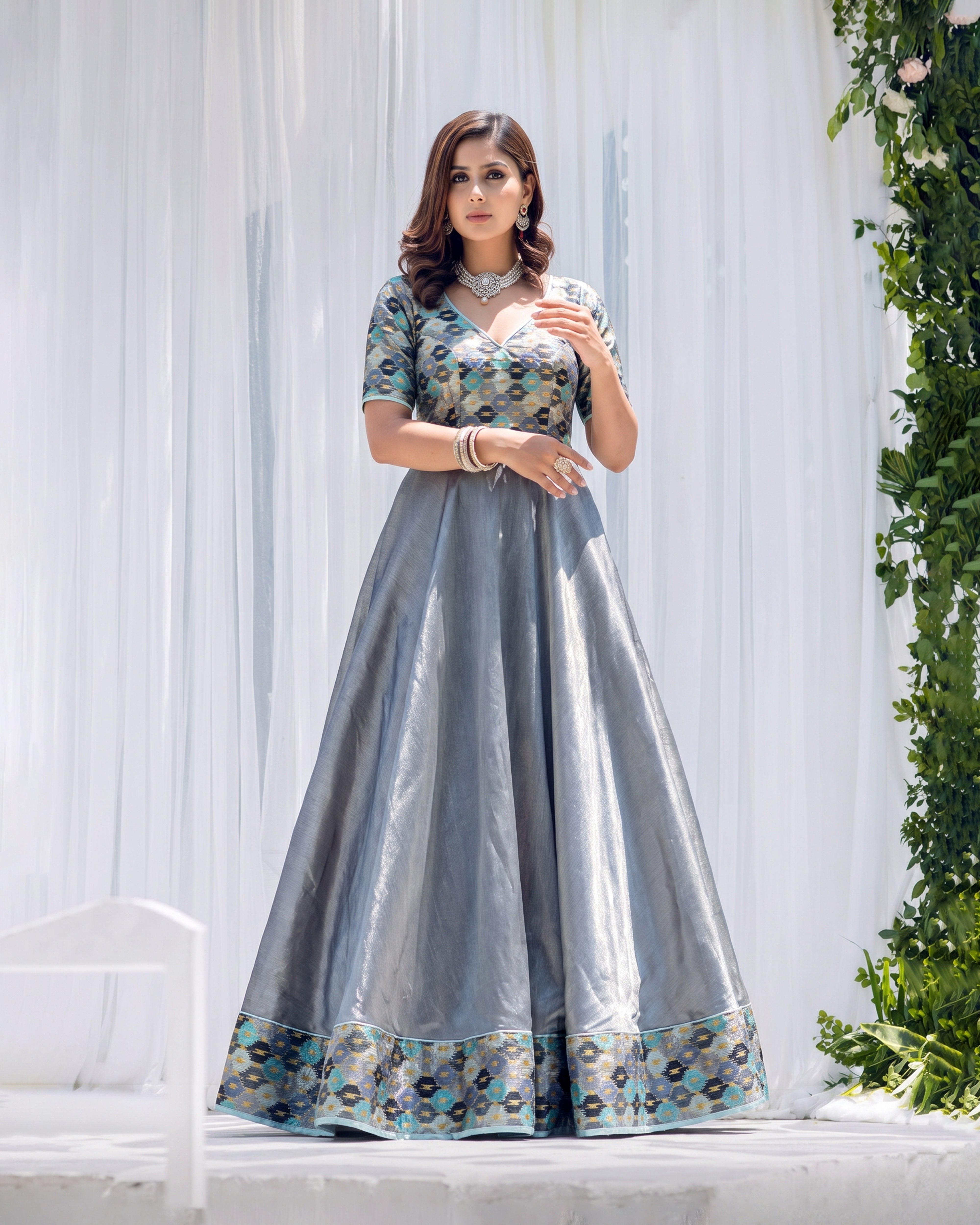 A woman models a floor-length gray anarkali gown with a heavily embroidered teal, gold, and white patterned bodice and border, set against a white draped backdrop with green foliage on the right.