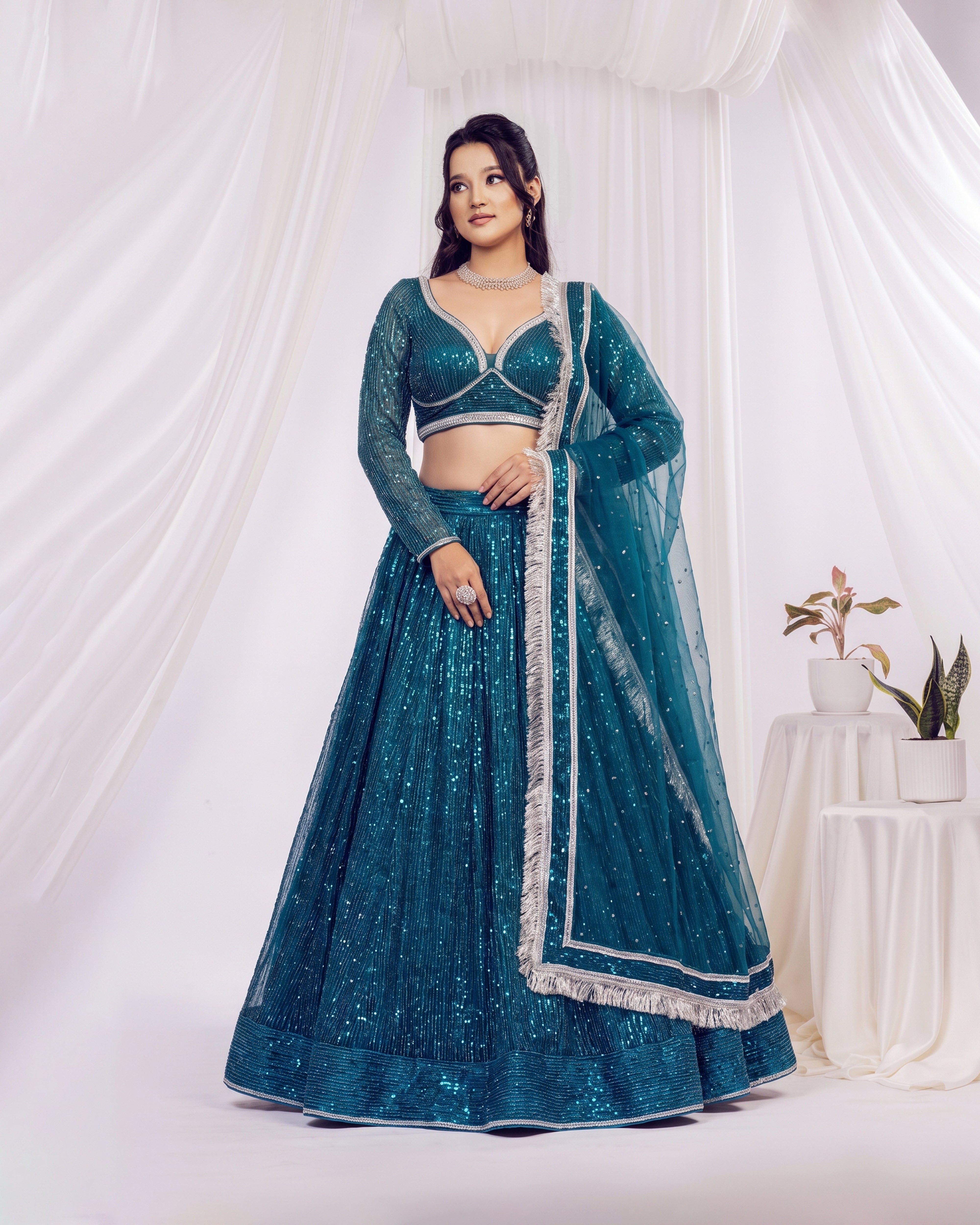 A woman models a teal blue sequined Indian ethnic outfit, consisting of a long-sleeve crop blouse, a flared skirt, and a matching sheer dupatta with silver borders, set against a draped white fabric backdrop.