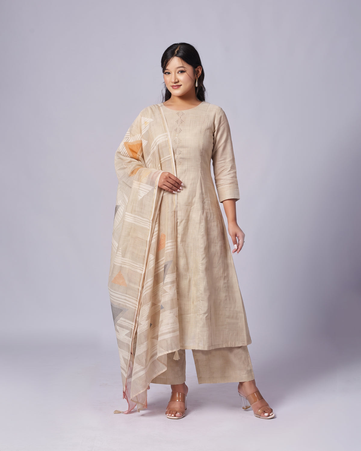 A woman modeling a beige three-piece traditional outfit with a matching patterned dupatta, standing against a light gray studio background.